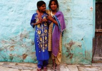 Two smiling Indian girls in red and purple saris stand in front of a turquoise wall