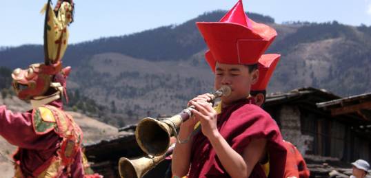 A monk from Bhutan playing a brass instrument and a brightly coloured dancer