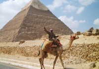 An Egyptian man riding his camel along the road, with a pyramid in the background