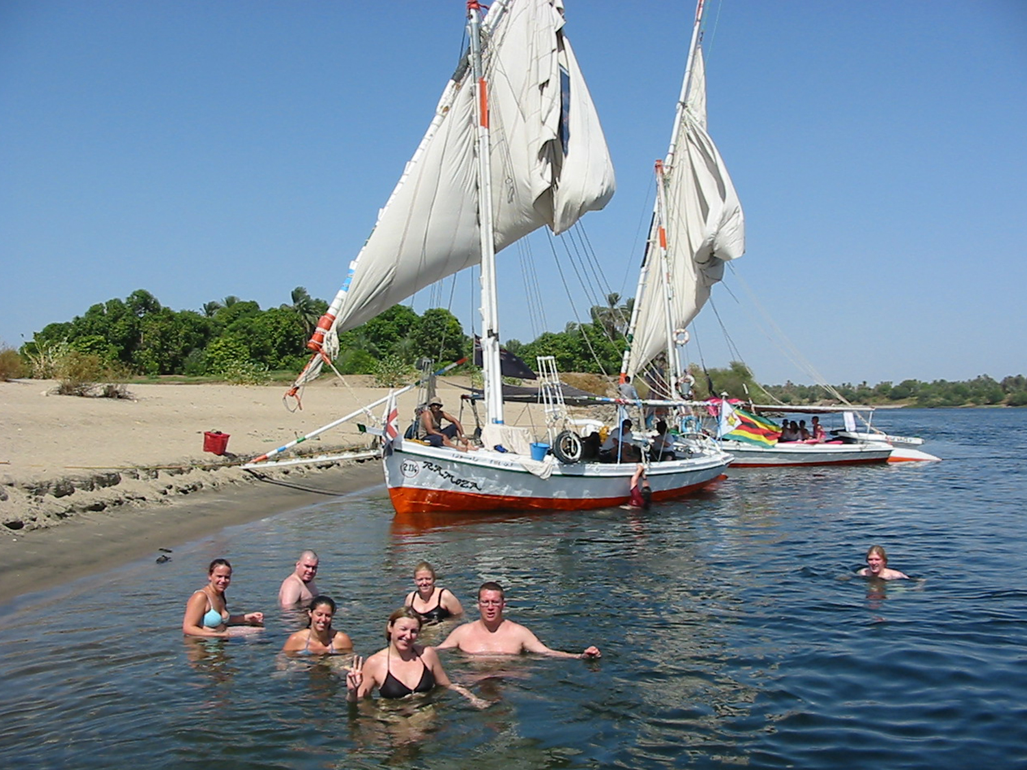 A felucca boat docked on the bank of the Nile with people enjoying swimming in the river.