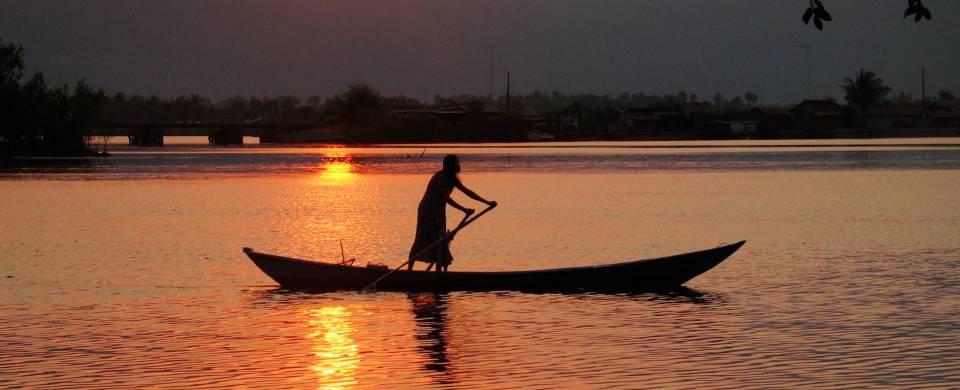 Long, thin boat being rowed across the water as the sun sets and stains the landscape orange in Kamp