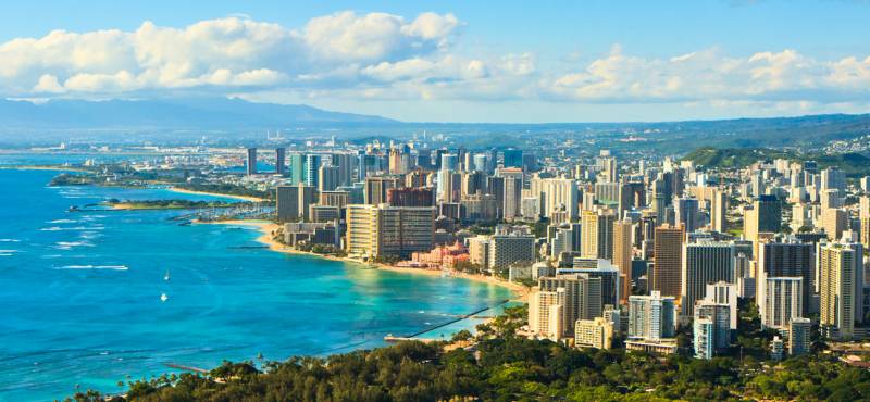 A panoramic view of the coastline of Oahu with skyscrapers fronting the beach and blue waters